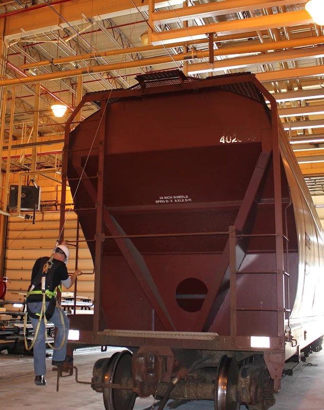 Worker in full fall arrest harness climbing a railcar ladder at an oil and gas processing plant