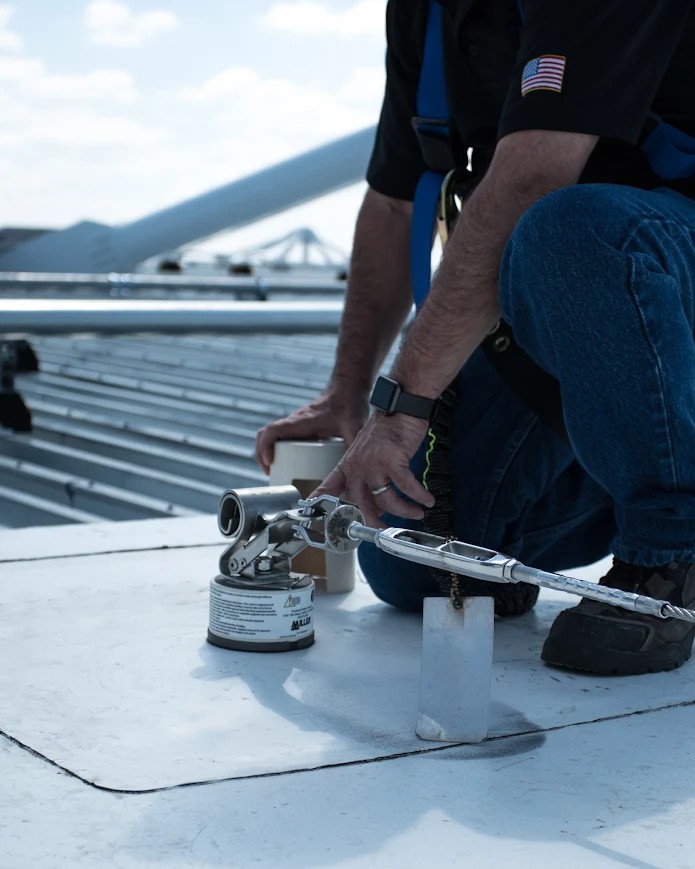 Worker installing a horizontal lifeline fall protection anchor on a metal rooftop at an oil and gas industrial site.